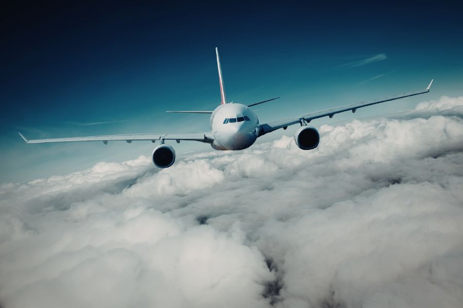 Un avion de ligne blanc survole un épais tapis de nuages blancs sous un ciel bleu, symbole d'un voyage de 3 semaines vers une destination lointaine.