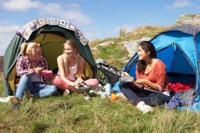 Trois jeunes femmes partagent un repas devant leurs tentes en pleine nature, une expérience authentique de trip à l'étranger.