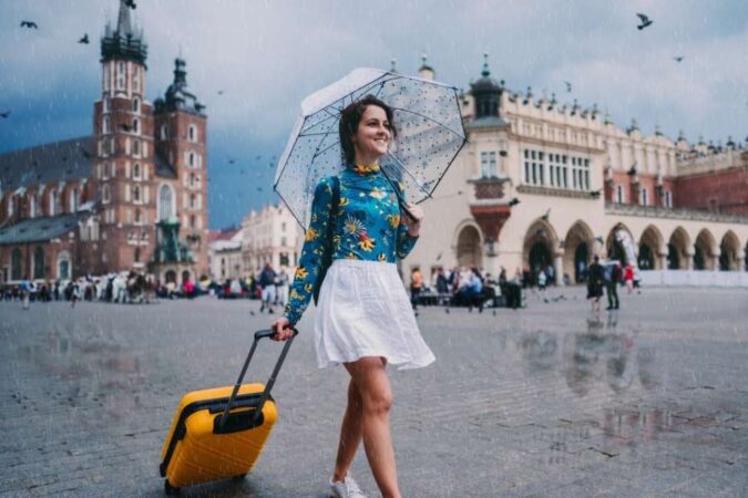 Une jeune femme souriante marche avec sa valise jaune et un parapluie sous une pluie légère lors d'un Voyage en solo sur une place historique.