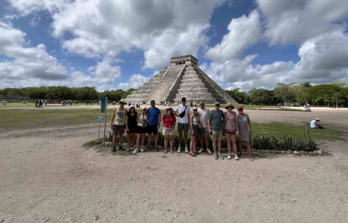 Un groupe de touristes pose pour une photo devant la grande pyramide d'El Castillo à Chichen Itza, Mexique, sous un ciel nuageux.