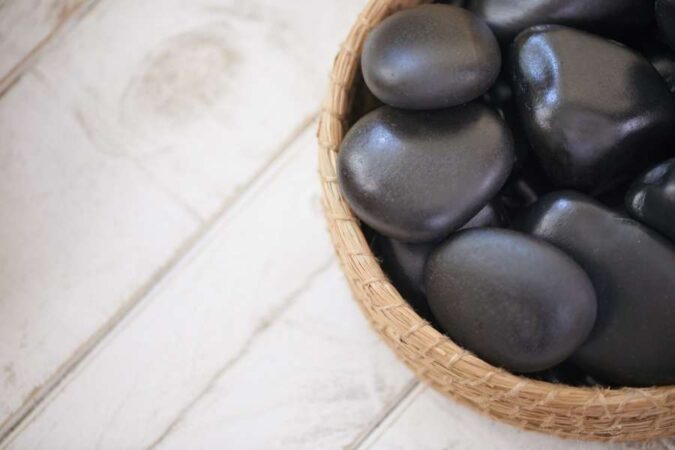 Un panier en osier rempli de pierres noires lisses et brillantes, prêtes pour une séance de massage aux pierres chaudes, est posé sur une table en bois clair.